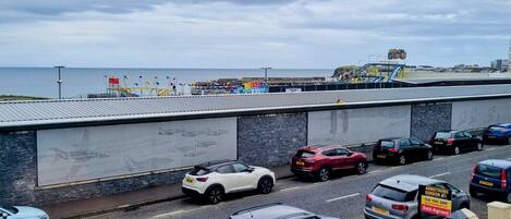 A parking area with multiple cars, a building with a stone facade, and a view of the sea in the background.