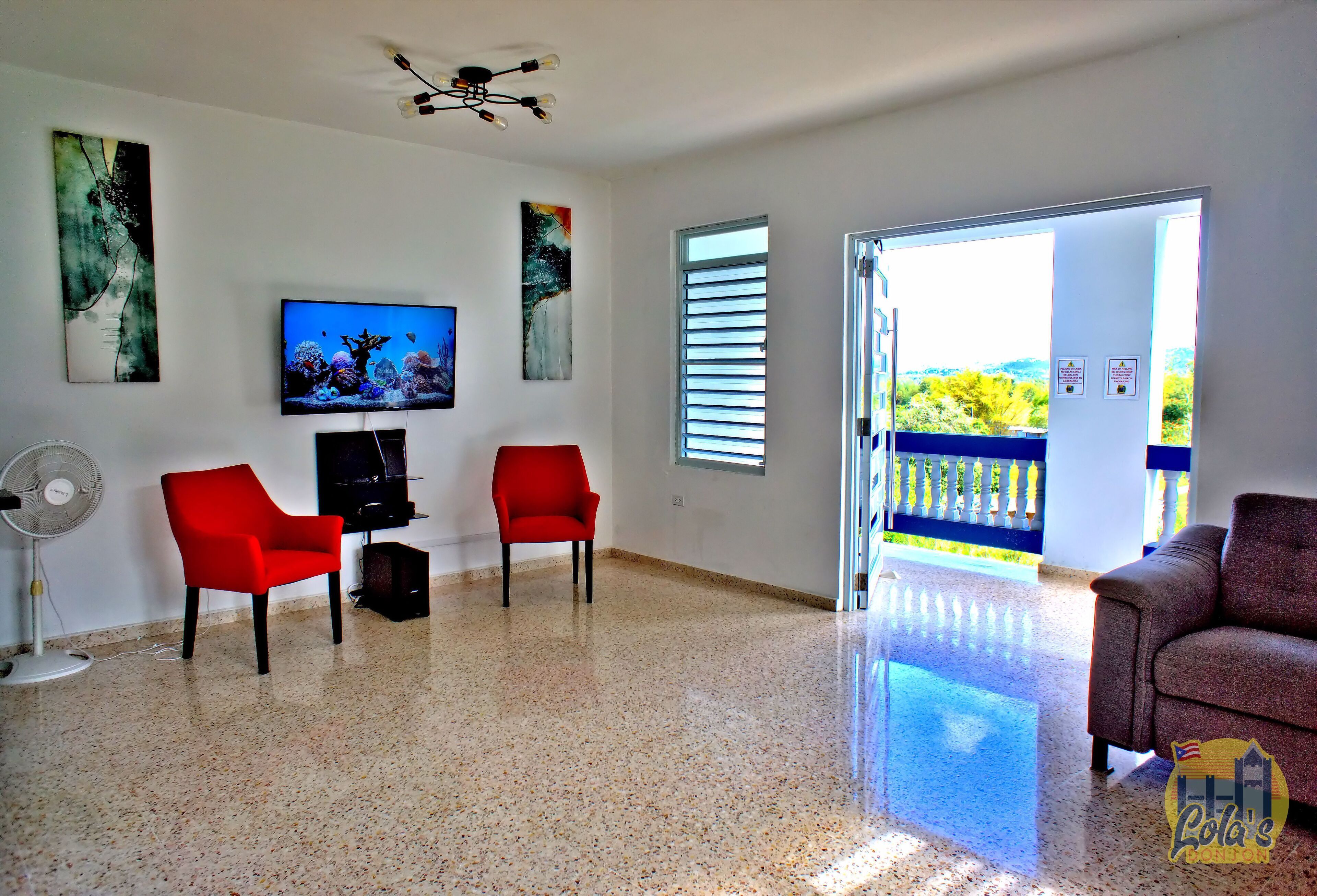 A spacious living room with a large TV, two red chairs, a fan, and a balcony with a view.