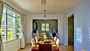 A dining area with a long table, chairs, and a vase with flowers. There are windows with blinds and a kitchen area in the background. - Lola's DonJon + Peaceful prop. + Near Beaches + HTUB (San German)