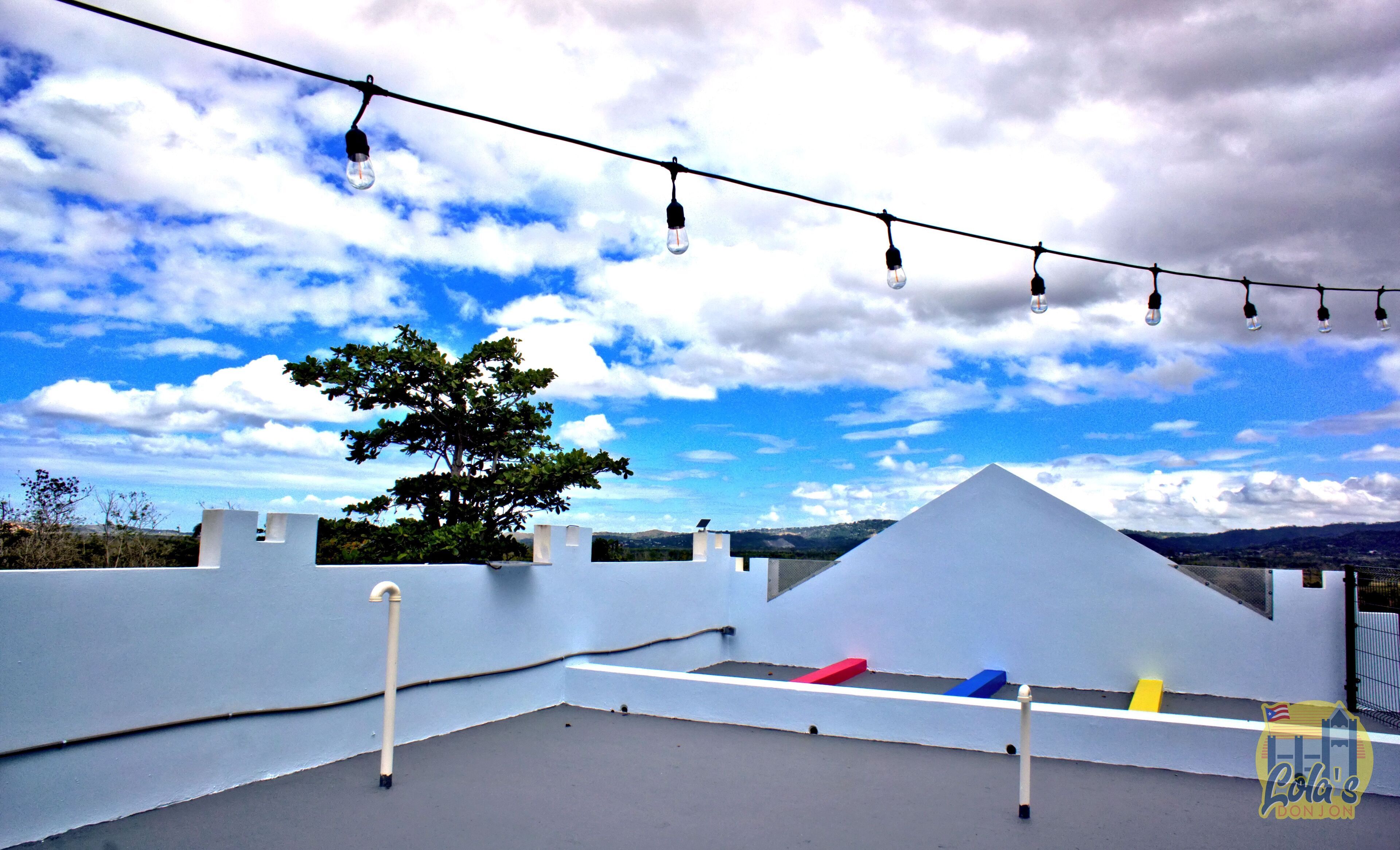 Una terraza con una estructura blanca, un árbol y una guirnalda de luces bajo un cielo parcialmente nublado.