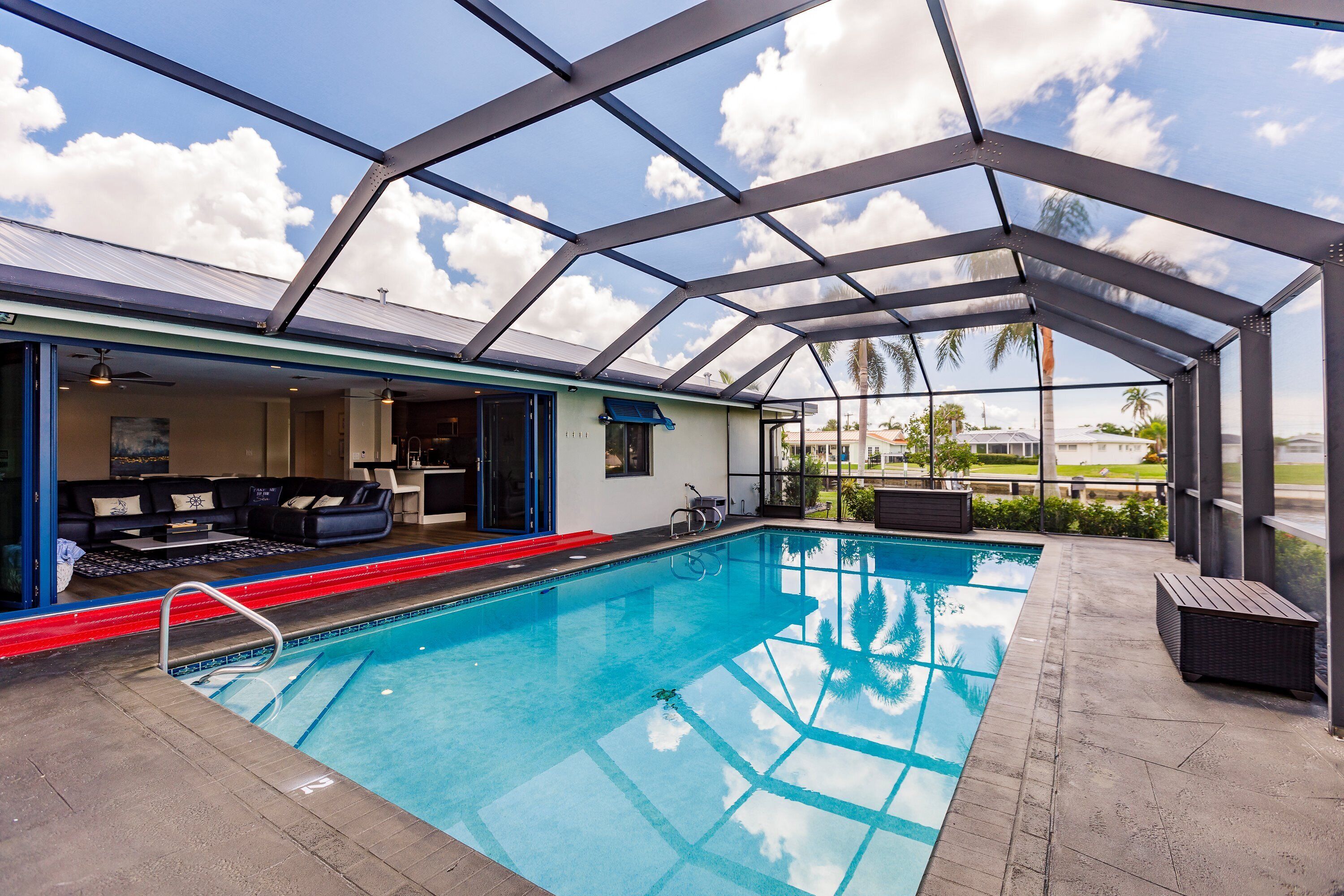 An indoor swimming pool with a glass ceiling, a lounge area with a sofa and coffee table, and a view of palm trees outside.