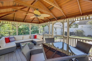 A covered outdoor seating area with wicker furniture, a glass table, and a ceiling fan.