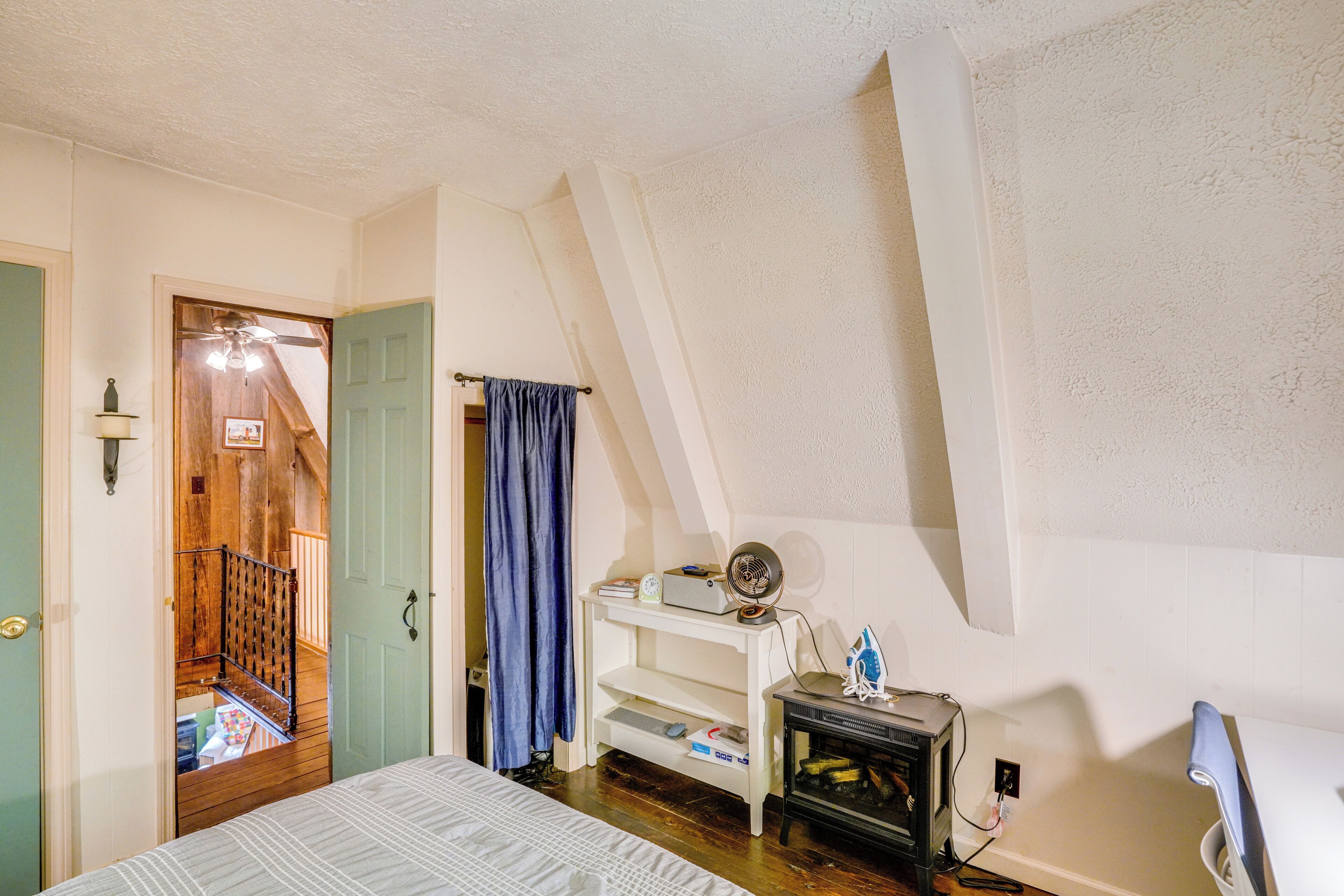 A bedroom with a bed, a small white shelf with a fan, a heater, and a door leading to a staircase.
