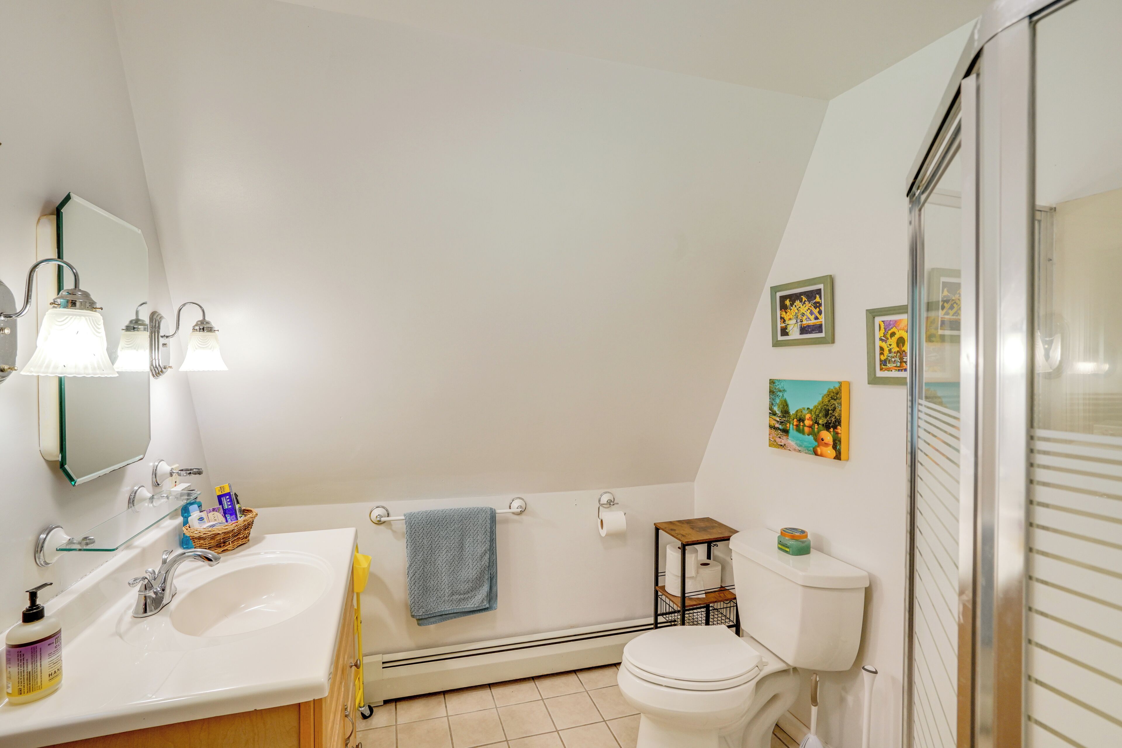 Un bagno con lavandino bianco, specchio e water. Due luci a parete, un asciugamano appeso al muro e una piccola mensola con articoli da toilette.