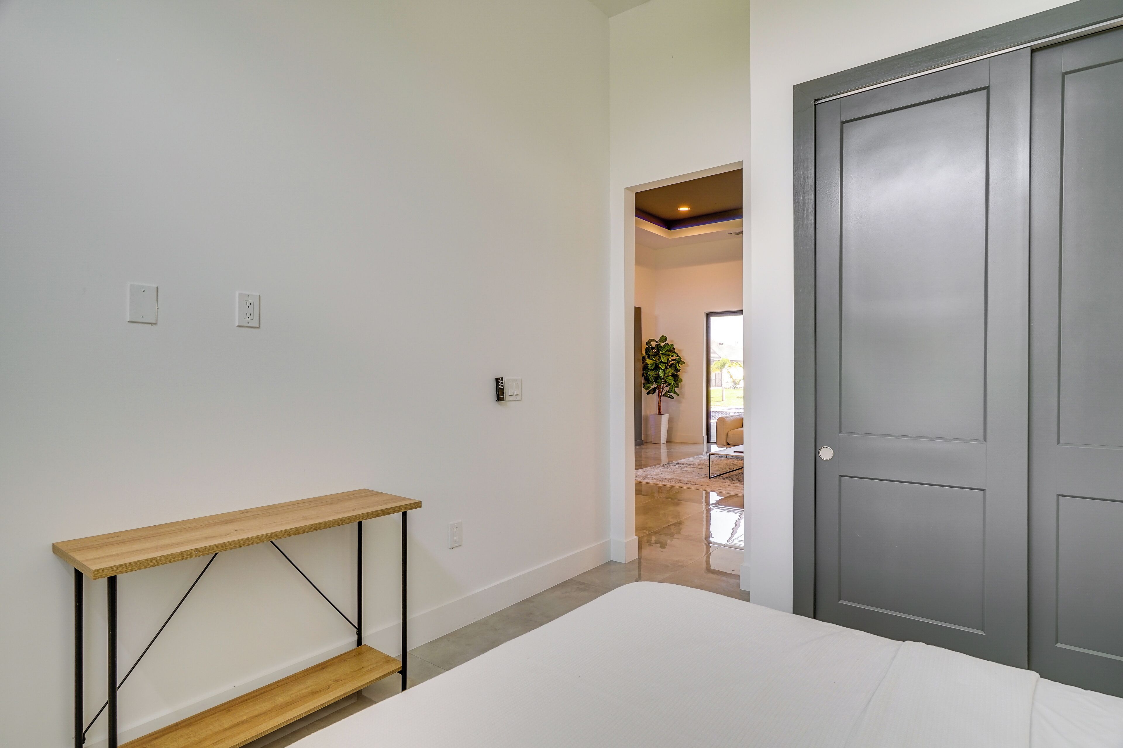 A bedroom with a bed, a wooden shelf, a gray door, and a hallway leading to another room.