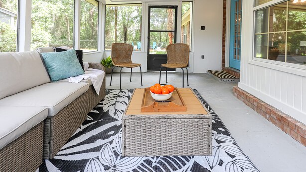 Une véranda couverte avec un canapé en osier, deux fauteuils, une table basse sur laquelle se trouve un saladier rempli de fruits, et un tapis à motifs.