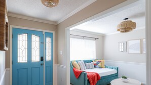 Une pièce avec une porte bleue, un canapé turquoise agrémenté de coussins, une table blanche et un luminaire suspendu.