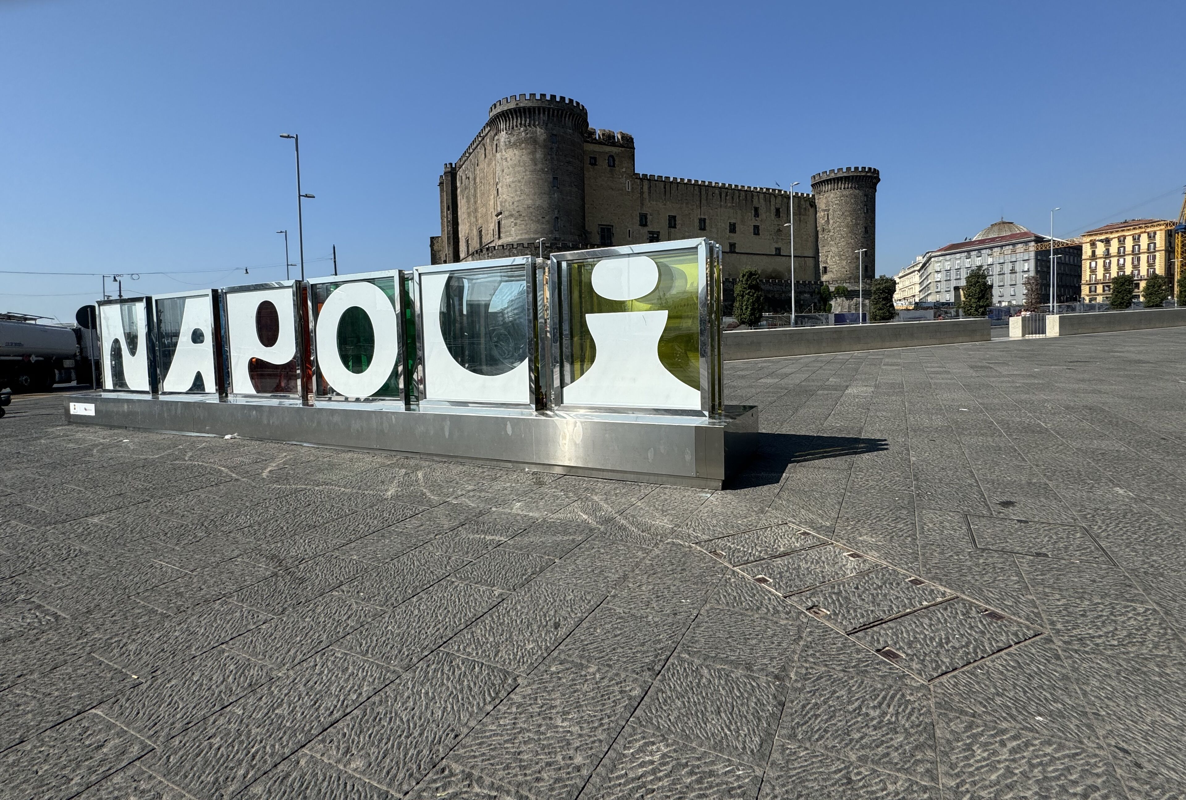 A large sign with the word "NAPOLI" in bold letters, set against a backdrop of historic buildings and a clear sky.