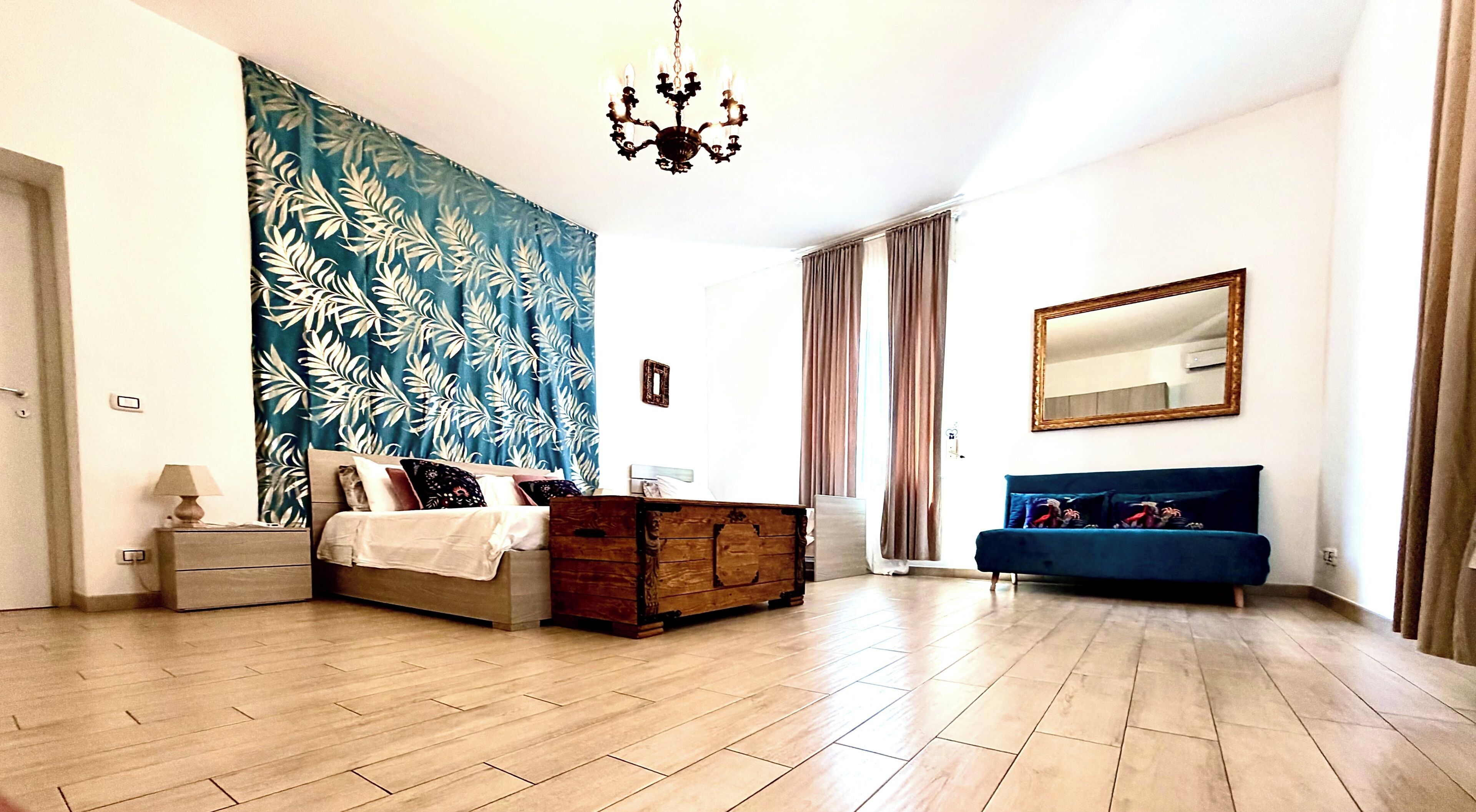 A modern bedroom with a large bed, a dresser, a blue sofa, a chandelier, and a large mirror.