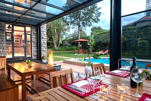 A dining area with wooden tables and chairs, a pool, and a view of the garden. - Deerpark Luxury Stays (Nairobi)