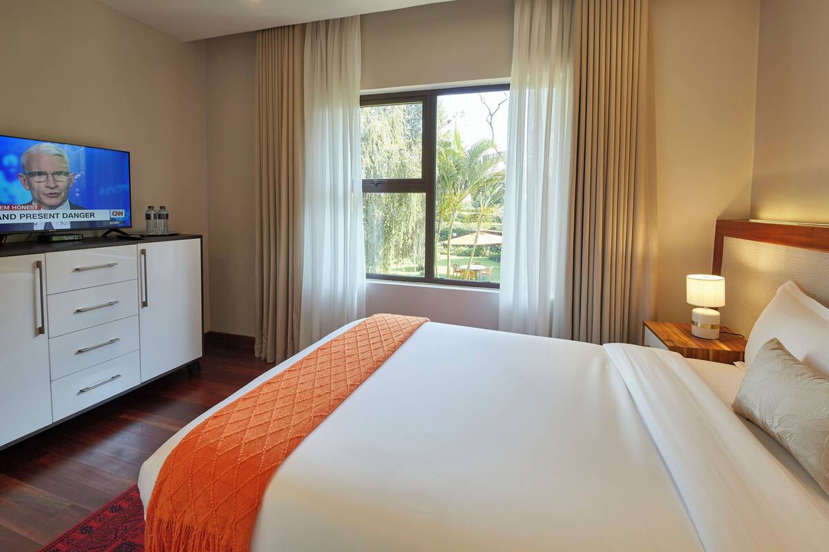Een hotelkamer met een bed, een televisie op een kast, een raam met gordijnen en een nachtkastje met een lamp.