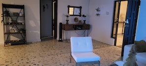 A living room with a white armchair, a wooden console table with decorative items, a mirror, and a shelf with plants.