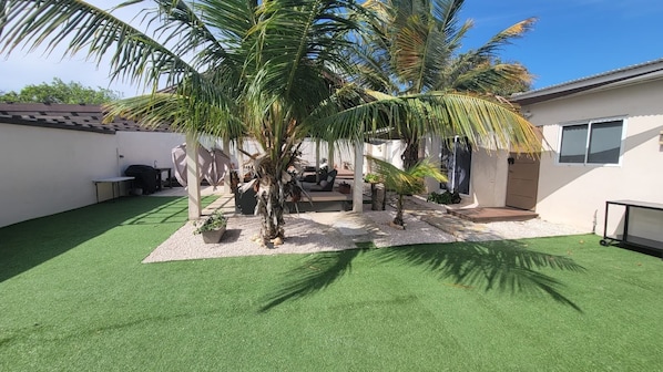 Un patio trasero residencial con césped artificial, palmeras, un área de patio y un edificio blanco con techo de tejas.