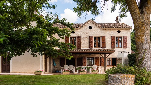 A two-story stone house with a covered patio, outdoor seating, and a large tree in the foreground. - Maison de Campagne "monplaisir" en Quercy Blanc (Montlauzun)