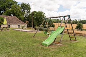 Un parque infantil con un tobogán verde y columpios, rodeado de césped y un edificio de piedra.