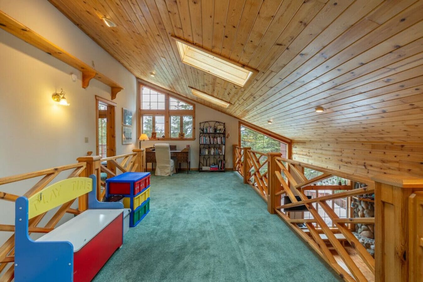 Une pièce mansardée spacieuse, avec un plafond en pente, des poutres en bois, un espace de jeu doté de bacs de rangement colorés, un bureau avec une chaise et une grande fenêtre offrant une vue sur l’extérieur.