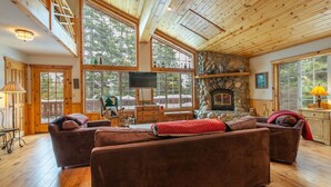 A cozy cabin interior with a stone fireplace, wooden ceiling, large windows, and a comfortable seating area. - Tahoe Donner w/ Private Hot Tub (Truckee)
