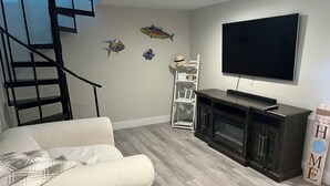 Ein Wohnzimmer mit einem weißen Sofa, einem an der Wand befestigten Fernseher, einer dunklen Unterhaltungseinheit und einer Treppe mit schwarzer Geländer.