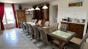 Comedor con una mesa larga, sillas y lámparas colgantes. Se observa también una zona de cocina con armarios y electrodomésticos.