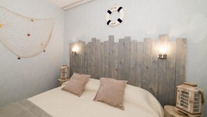 Un dormitorio con cama, dos almohadas, cabecero de madera, dos luces fijadas a la pared, un adorno de círculo salvavidas y una pequeña mesa de madera con un farol.