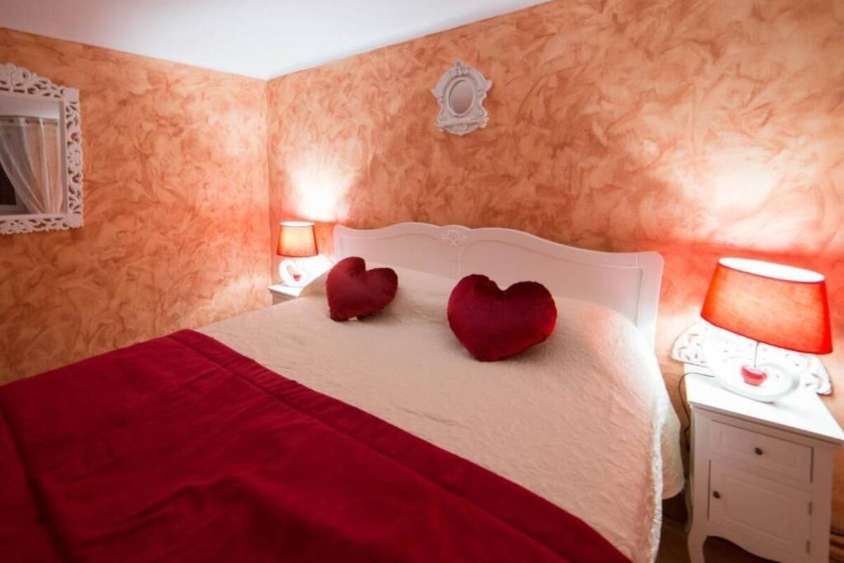 Ein Schlafzimmer mit einem Bett, zwei herzförmigen Kissen, einer roten Decke, einem Nachttisch mit Lampe und einem dekorativen Spiegel an der Wand.