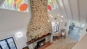 Uma sala de estar ampla com lareira de pedra, piso de madeira e grandes janelas com obras de arte coloridas.