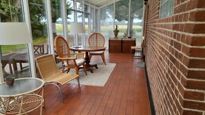 A covered patio with wicker furniture, a brick wall, and large windows overlooking a field.