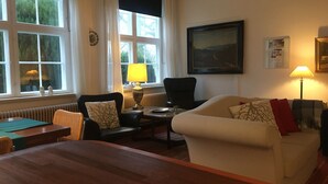 Un salon chaleureux avec une table basse en bois, un canapé blanc et un fauteuil noir. Il y a deux fenêtres avec des rideaux, un tableau au mur et une lampe sur une table d’appoint.