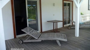 Un espacio al aire libre techado con una terraza de madera, una silla de descanso, una mesa y un ventanal corredero que da a una piscina.