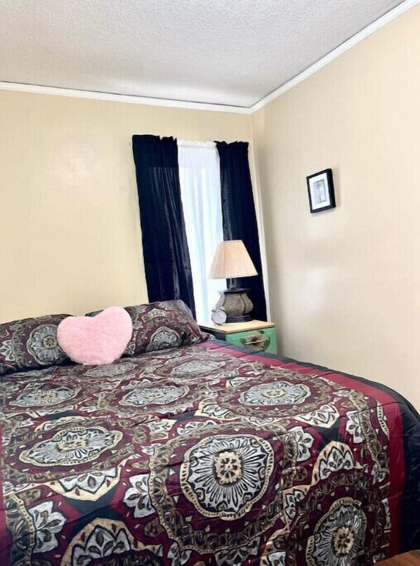 Een slaapkamer met een bedovertrek met patroon, een hartvormig kussen, een nachtkastje met een lamp en een ingelijst schilderij aan de muur.