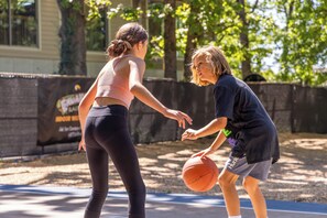 Dos personas jugando baloncesto en una cancha al aire libre, con árboles y un edificio al fondo.