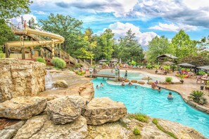 A pool area with multiple water features, including a waterfall and a slide, surrounded by rocks and greenery. - Lakeside Resort Cabin Steps to Table Rock Lake - Minutes to SDC! (Branson)