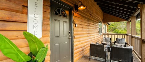 Une cabane en bois avec une pancarte « BIENVENUE », une porte grise, des meubles en osier et une véranda offrant une vue imprenable.