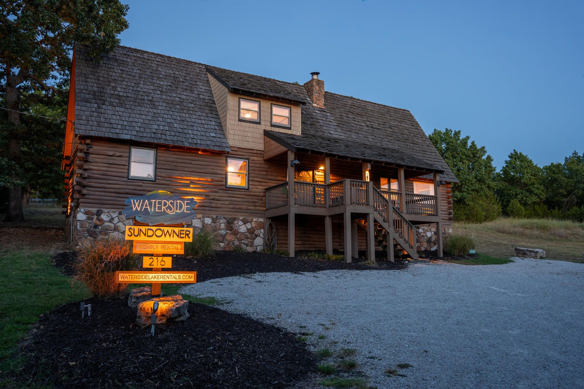 A log cabin with a stone foundation, a wooden deck, and a staircase leading to a second-story balcony.
