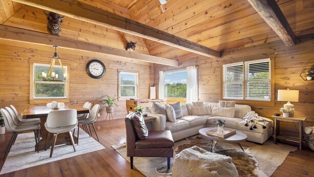A cozy cabin interior with a wooden ceiling, a dining area with a table and chairs, a living room with a sofa and a coffee table, and a large window with blinds. - Lakeside Resort Cabin Steps to Table Rock Lake - Minutes to SDC! (Branson)