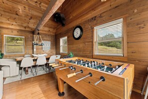 A wooden-floored room with a foosball table, a clock, and a dining area with chairs and a table. - Lakeside Resort Cabin Steps to Table Rock Lake - Minutes to SDC! (Branson)