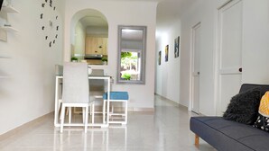 Un soggiorno moderno con un divano, un'area pranzo con sedie, una cucina visibile tramite un arco e un corridoio che conduce a una porta.