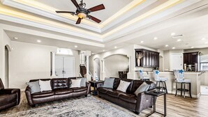A spacious living room with a large brown leather sofa set, a coffee table, and a patterned rug. There is a ceiling fan, recessed lighting, and a kitchen area with dark cabinets and a white countertop.