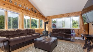 Ein gemütliches Wohnzimmer mit einem großen Ecksofa, einem passenden Hocker und einem gemusterten Teppich. Mehrere Fenster bieten einen Blick ins Freie.