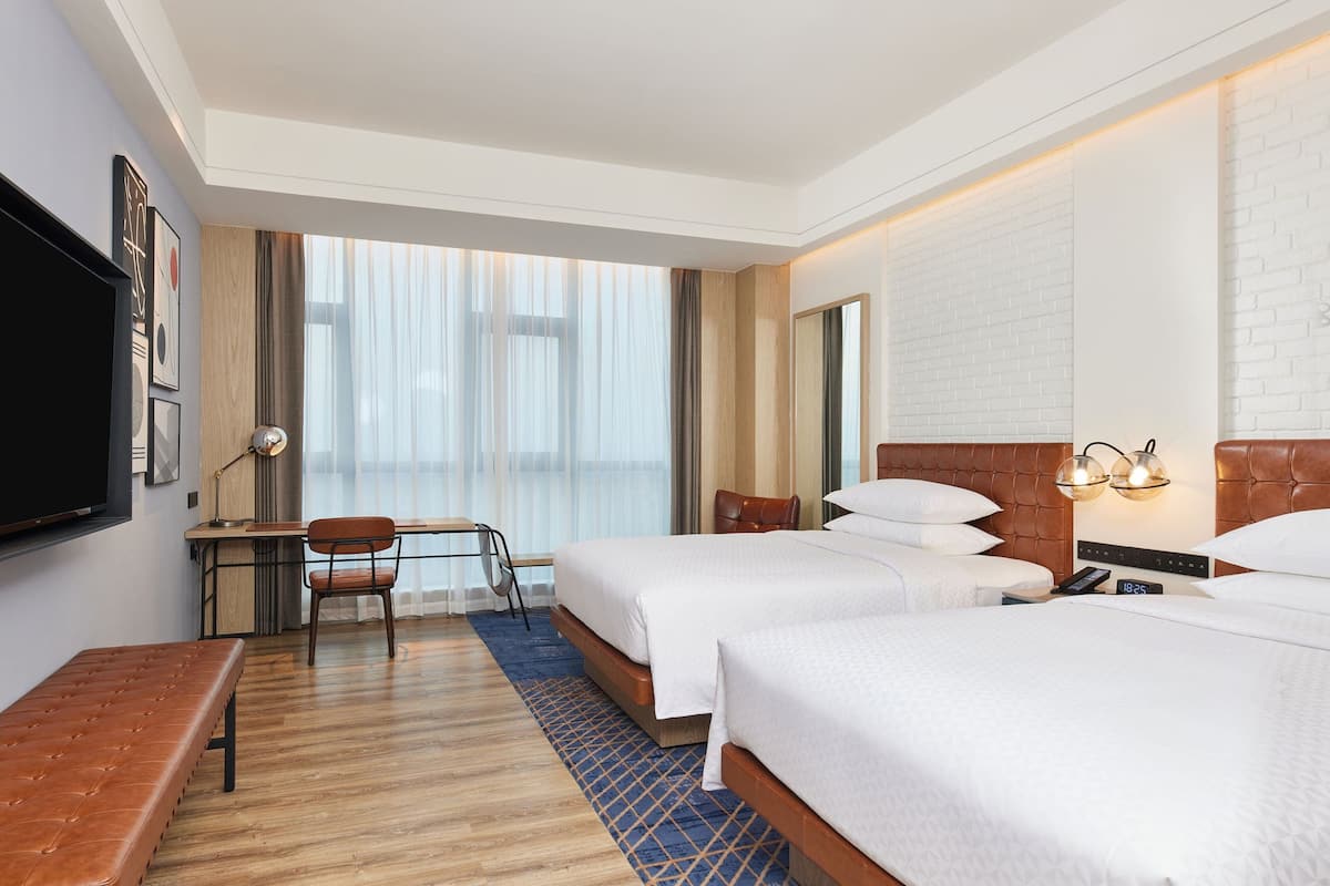 A hotel room with two beds, a desk, a chair, a TV, and a large window with curtains.