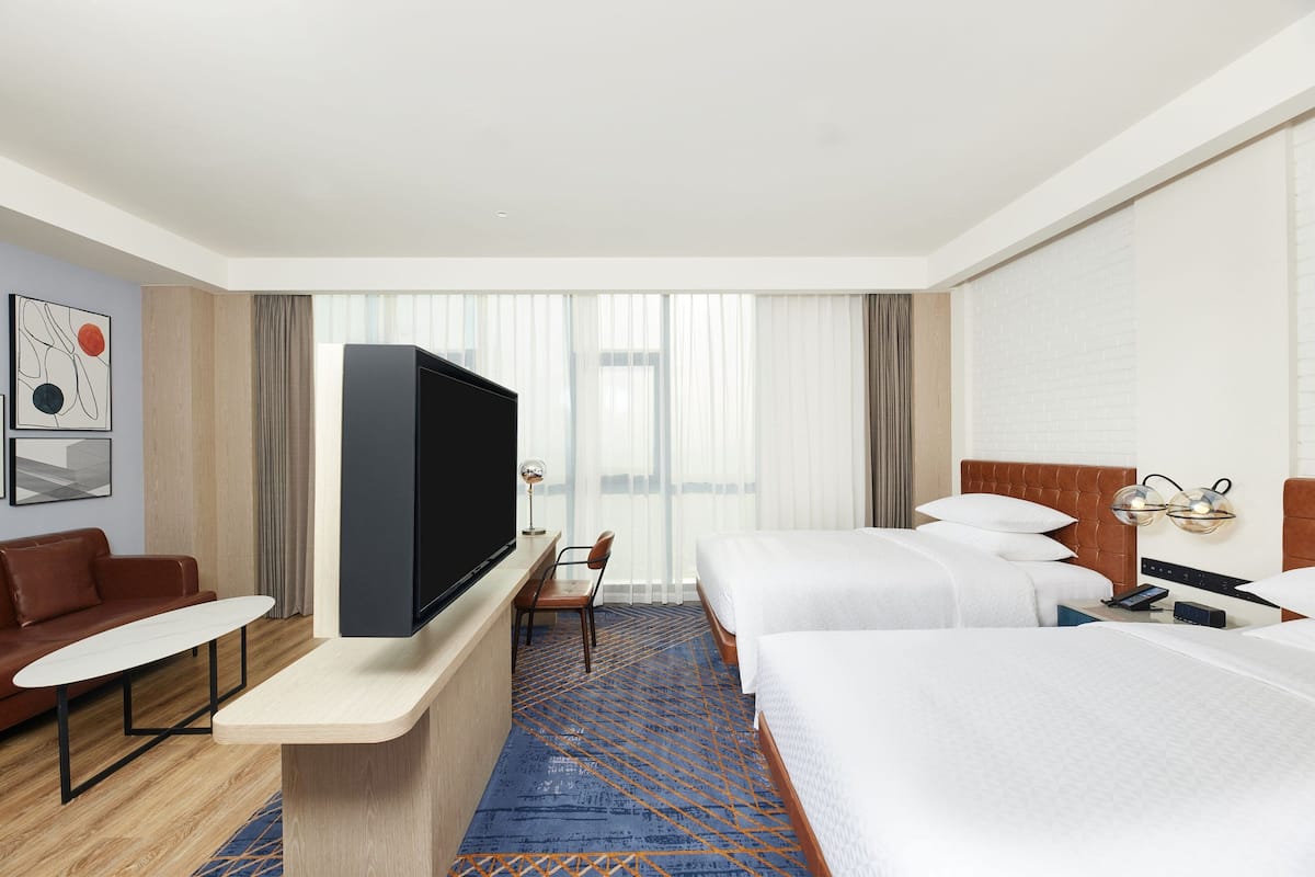 A hotel room with a flat-screen TV, two beds, a sofa, a small table, and a large window with curtains.