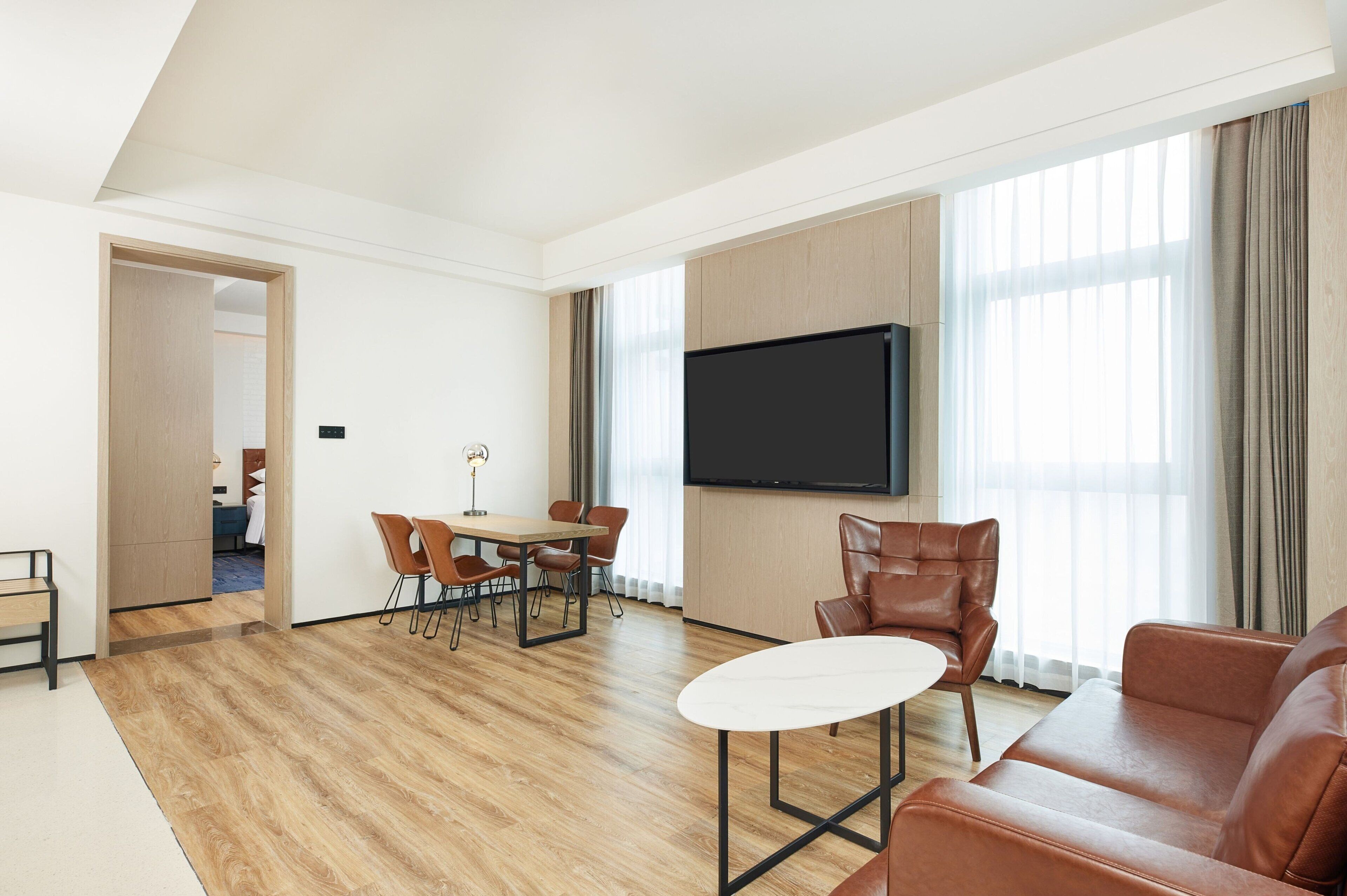 Un salon moderne avec un plancher en bois, une table ronde blanche, des canapés en cuir marron, un téléviseur fixé au mur et de grandes fenêtres avec des rideaux.