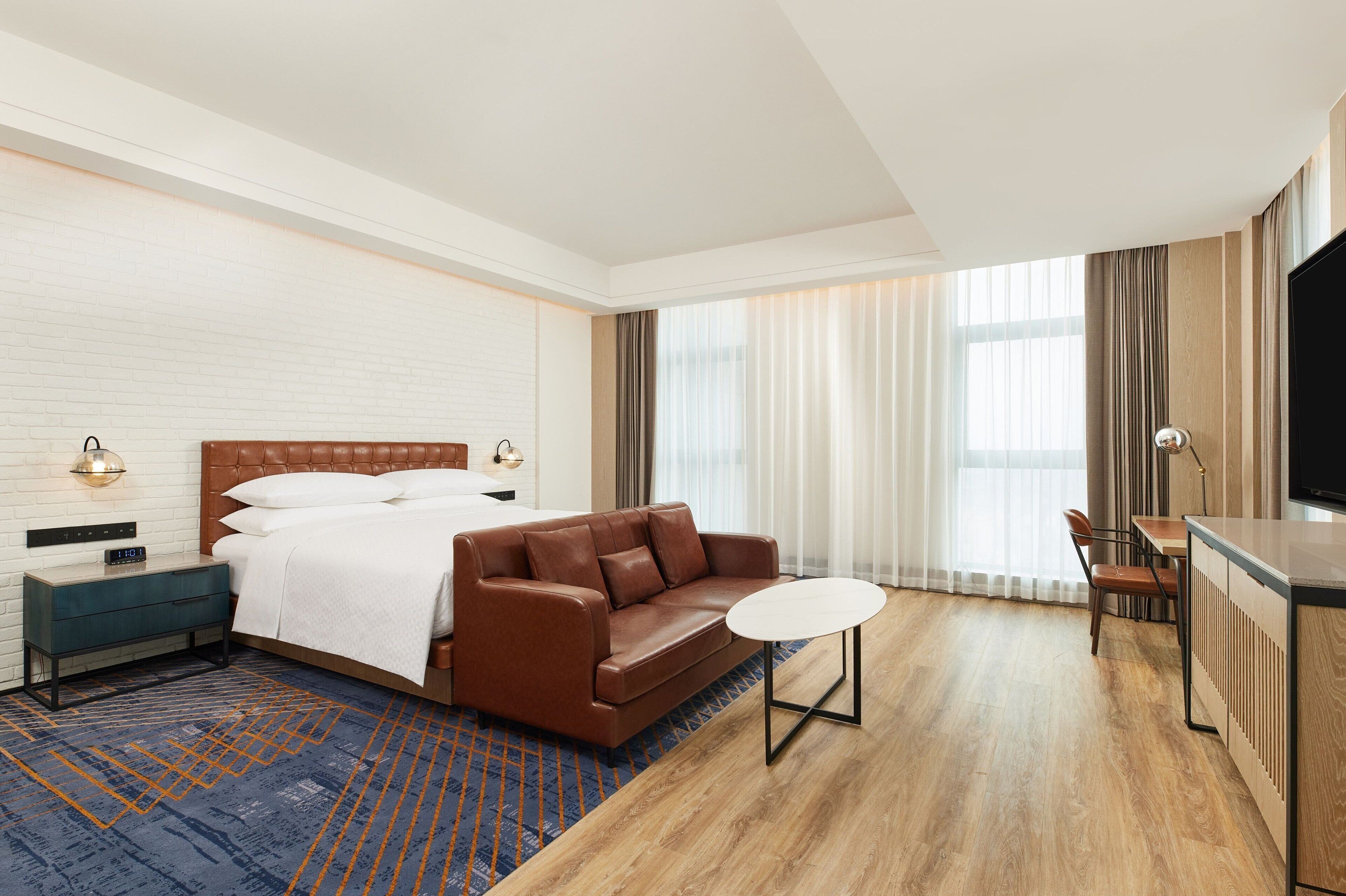 A hotel room with a bed, a brown leather sofa, a small round table, a TV, and a desk with a chair.