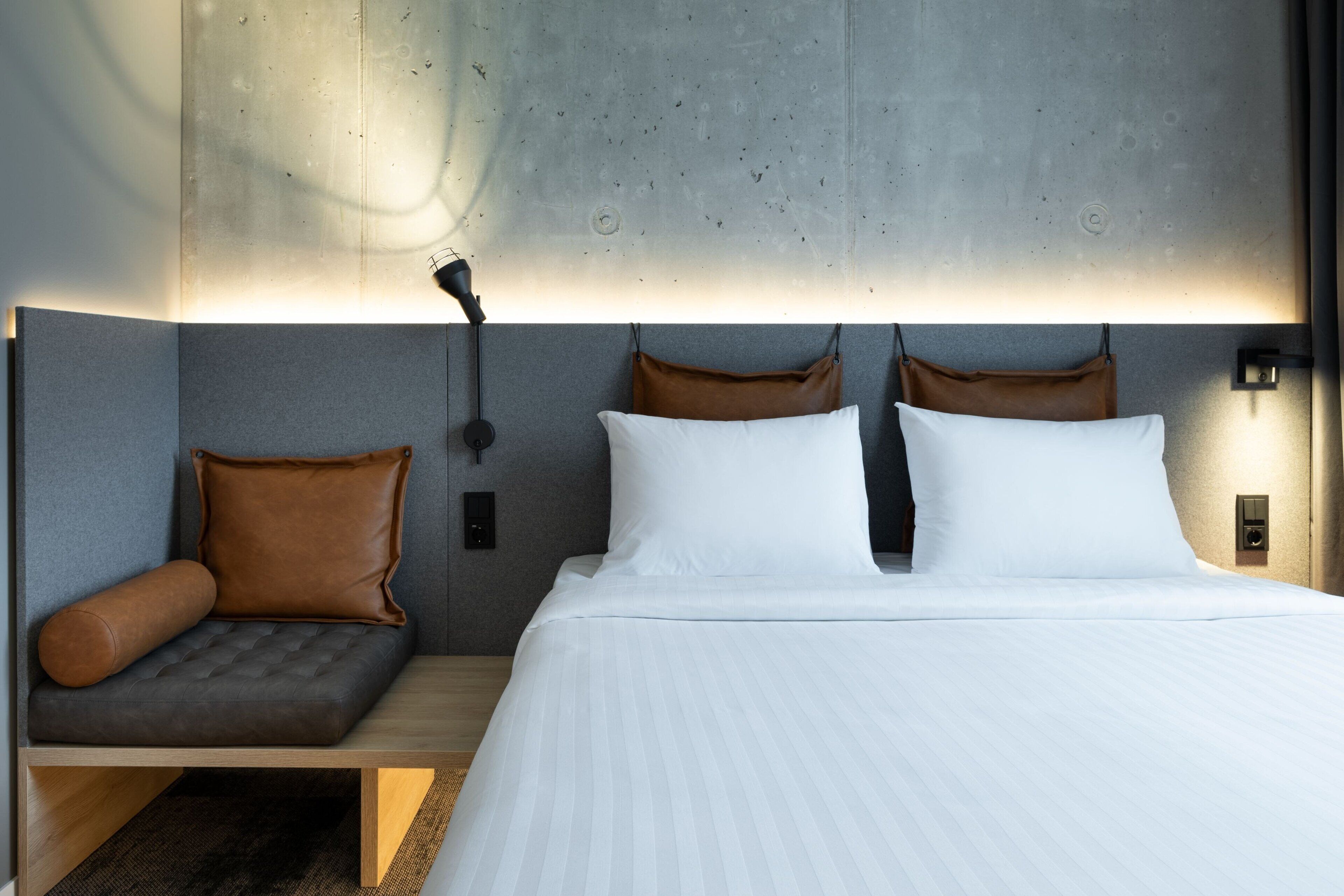 A modern hotel room with a large bed, a seating area with a cushioned bench, and a wall-mounted light fixture.
