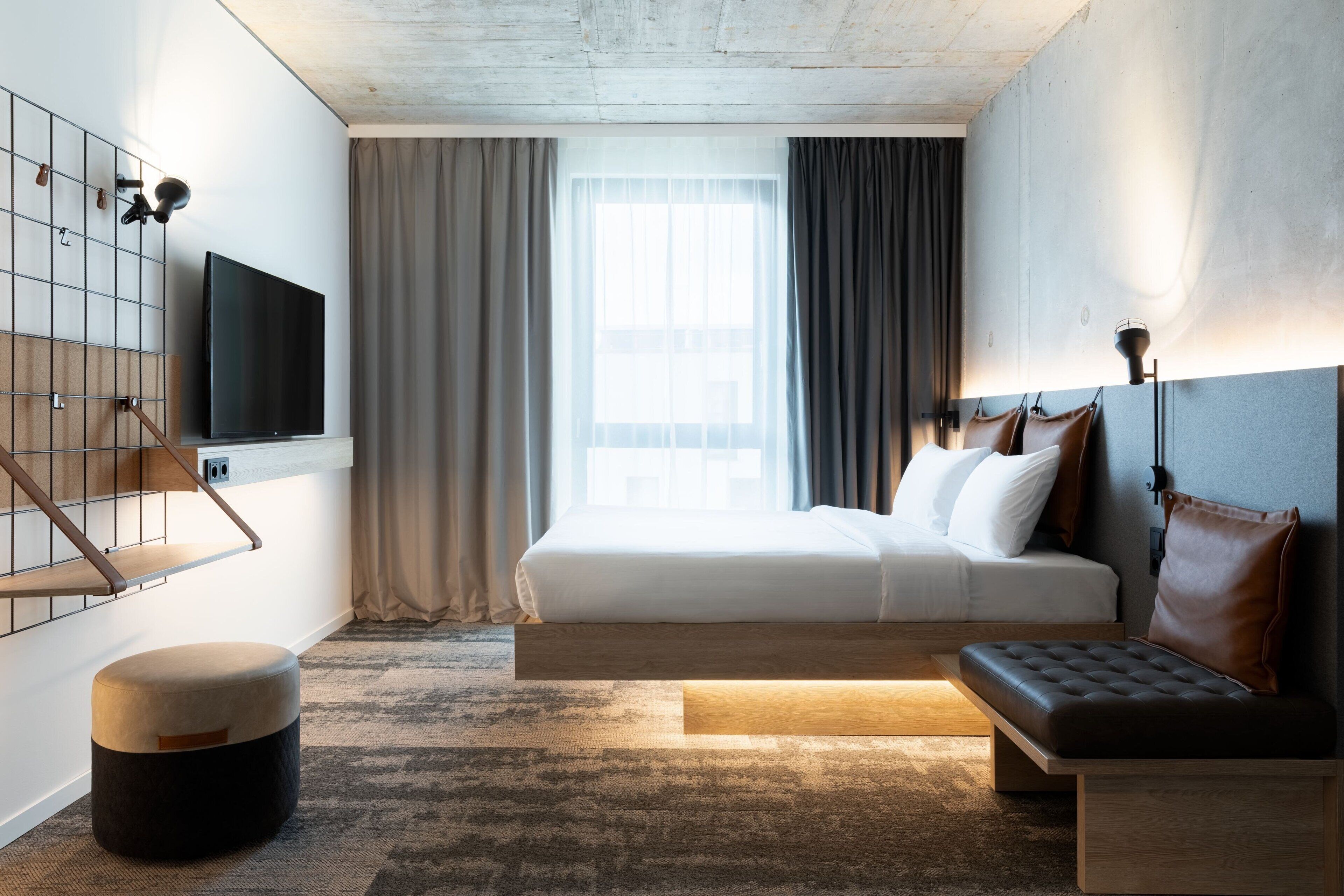 Una habitación de hotel moderna con una cama grande, un televisor montado en la pared, un banco de madera y un pequeño otoman.