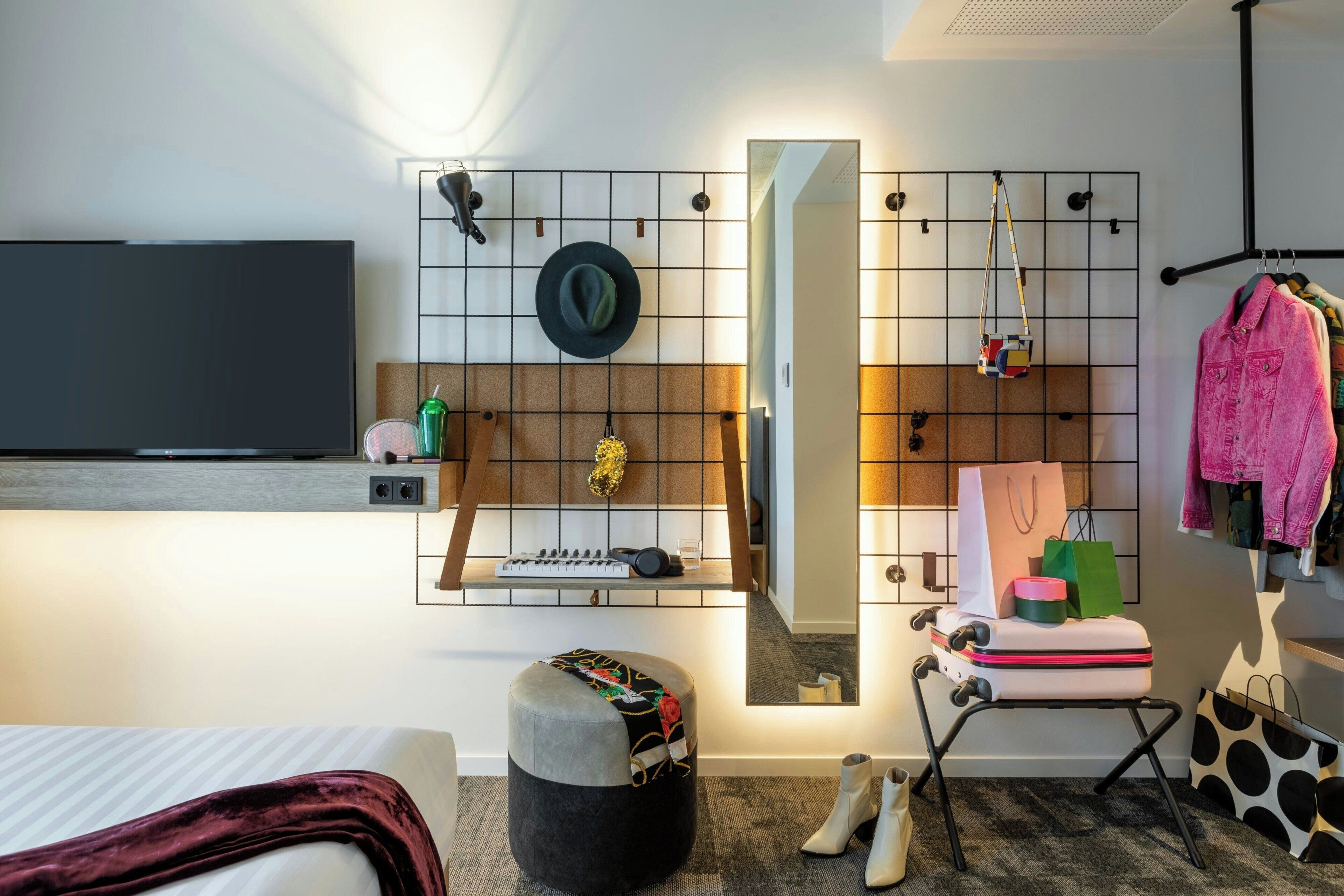 A modern hotel room with a large flat-screen TV, a mirror, a seating area with a chair and ottoman, and a wardrobe with hanging clothes.