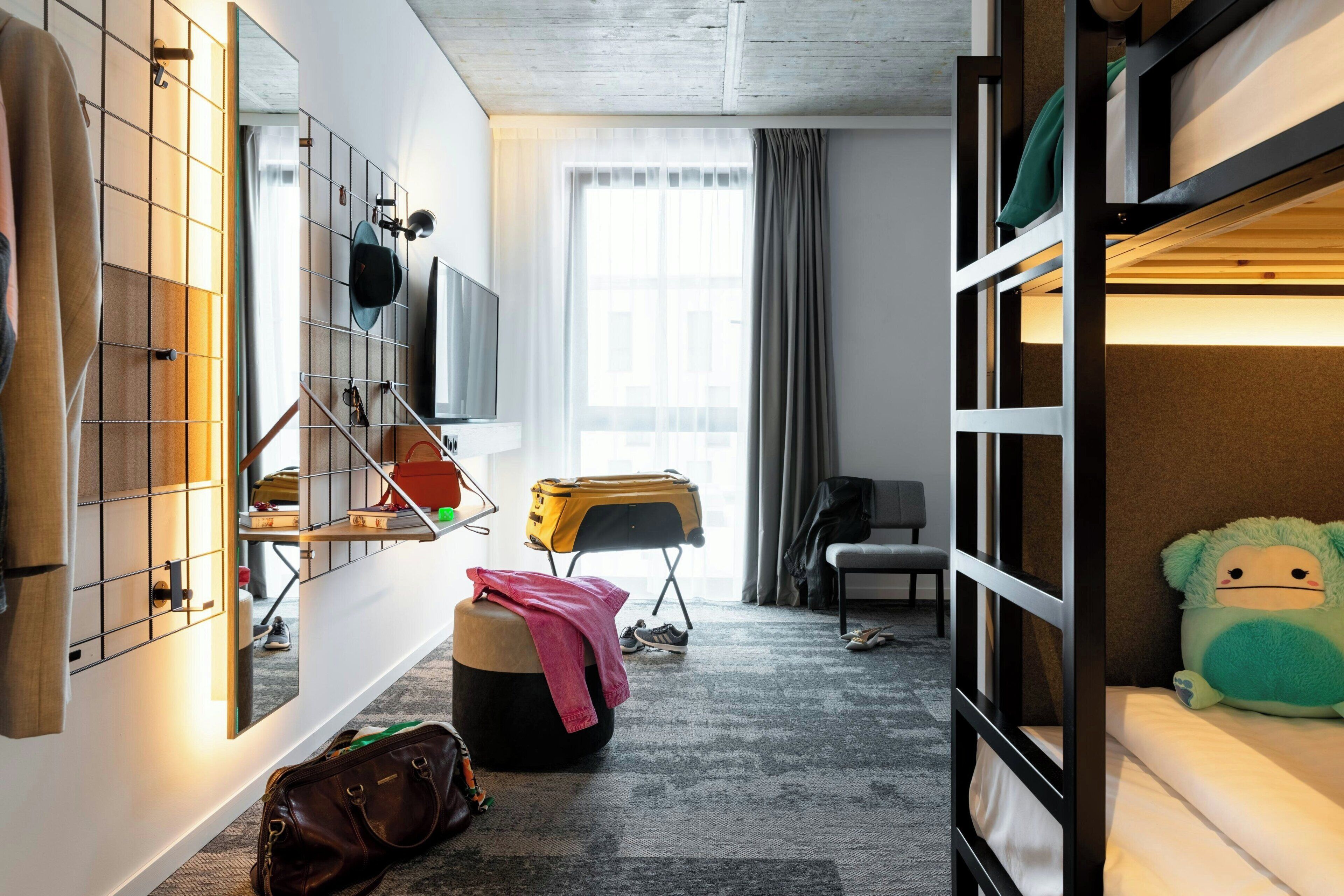 A room with bunk beds, a desk with a chair, a mirror, a window with curtains, and a suitcase.