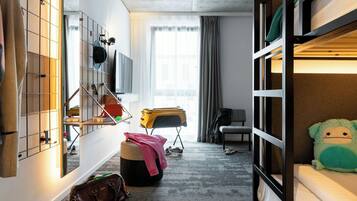 Una habitación con literas, un escritorio con silla, un espejo, una ventana con cortinas y una maleta.