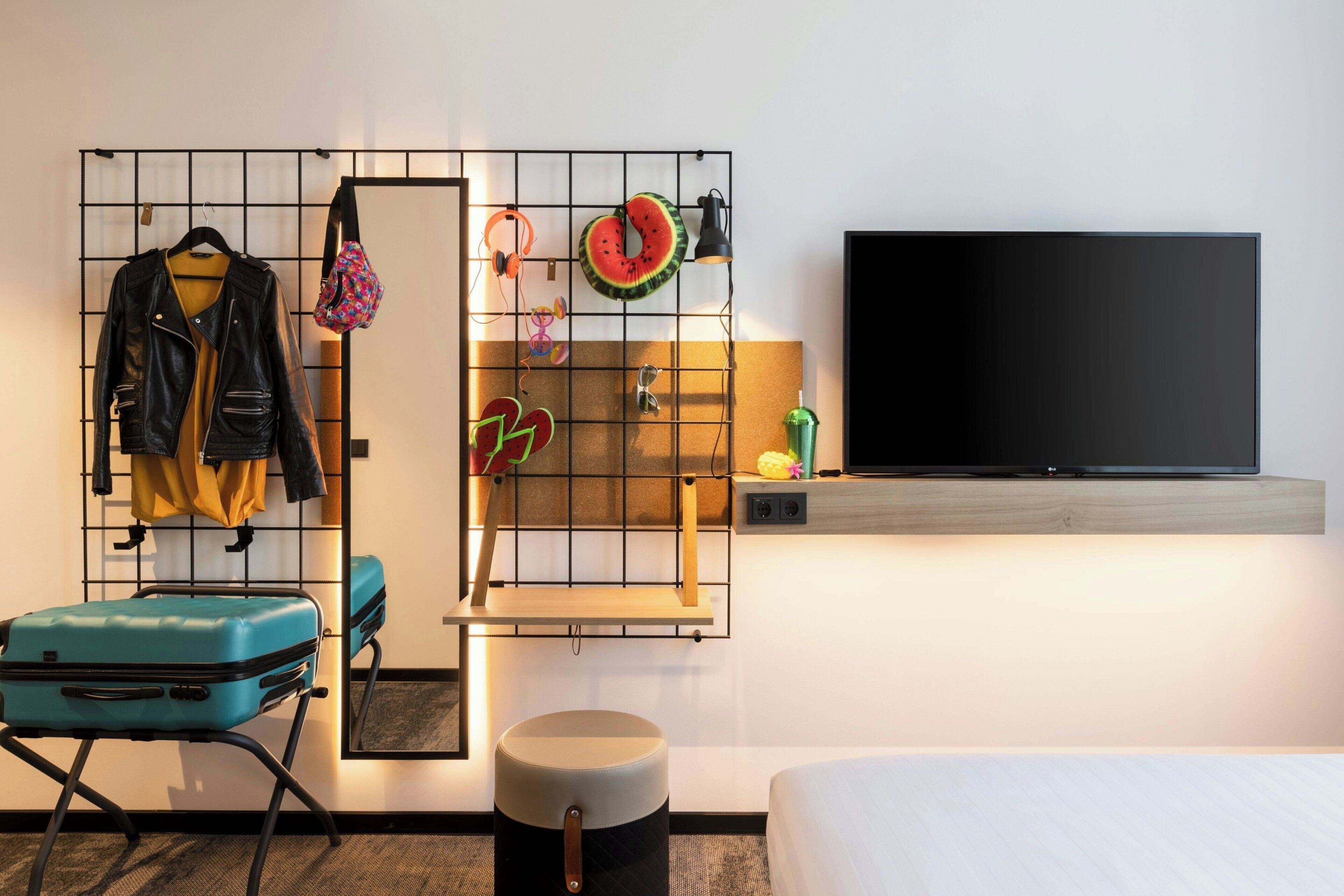 A hotel room with a bed, a TV, a suitcase, a mirror, and a wall-mounted shelf with various items.