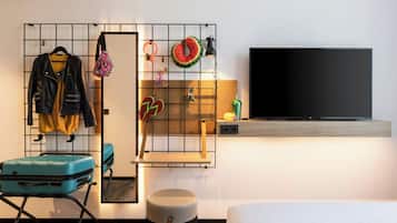 A hotel room with a bed, a TV, a suitcase, a mirror, and a wall-mounted shelf with various items.